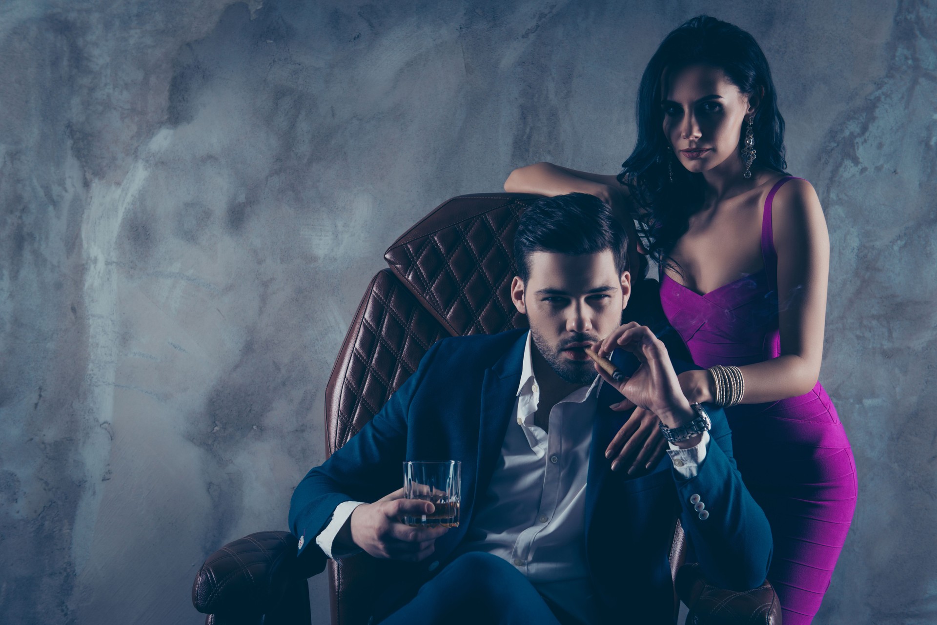 Portrait of brutal gentleman in formal wear sitting in leather chair holding glass with whiskey cigar in hands, charming pretty lady standing near looking at camera isolated on grey background